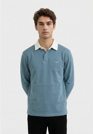 Ung mann med brunt hår iført en blå langermet polotrøye med hvit krage, stående mot en enkel lys bakgrunn.