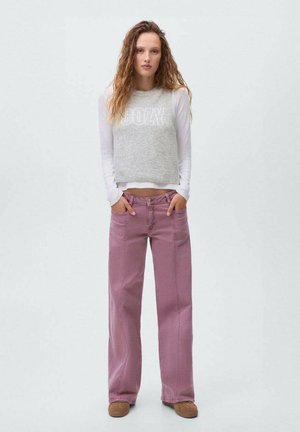Siv pulover s kratkimi rokavi z napisom "COZY", v kombinaciji s širokimi roza kavbojkami. Model stoji z rokama v žepih, obut v rjave čevlje.