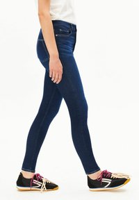 Dunkelblaue Denim-Jeans mit schmaler Passform, seitlichen Taschen und Steppnähten, kombiniert mit schwarzen Sportschuhen mit Gummisohle.