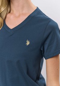 Tumši zila kokvilnas T-krekls ar V veida izgriezumu un īsām piedurknēm, uz kreisā krūtīm neliela zelta izšūta logo. Gluda tekstūra.