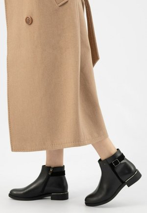 Zwarte leren enkellaarsjes met een zijrits, zilveren accenten en een gehelst zwarte band aan de bovenkant. Draagt een beige lange jas.