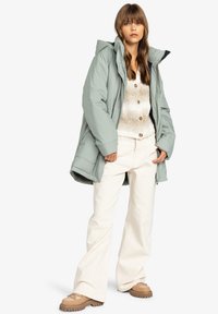 Jachetă puffer verde deschis cu glugă, asociată cu un pulover din tricot bej și pantaloni albi evazați. Încălțămintea este de culoare bej cu o talpă texturată.