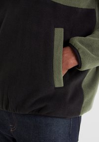 Fleece pullover in dark green and black, featuring a front pocket, textured fabric, and elastic hem. Paired with denim jeans.