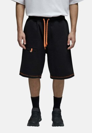 Homme portant un short noir avec des cordons et des coutures orange, des chaussettes noires, des chaussures noires et un t-shirt blanc, debout sur un fond blanc.