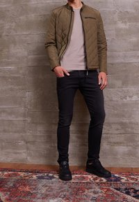 Quiltad olivgrön jacka med ståkrage, ficka med dragkedja, beige t-shirt, svarta slim fit-jeans och svarta sneakers, stående på en mönstrad matta.