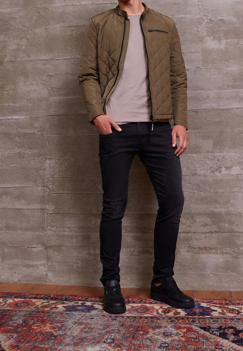 Quiltad olivgrön jacka med ståkrage, ficka med dragkedja, beige t-shirt, svarta slim fit-jeans och svarta sneakers, stående på en mönstrad matta.