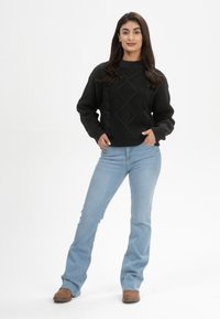 Schwarzer Strickpullover mit einem strukturierten Muster, ausgestattet mit einem Rundhalsausschnitt und ribbeligen Bündchen, kombiniert mit hellblauen Schlagjeans und braunen Schuhen.