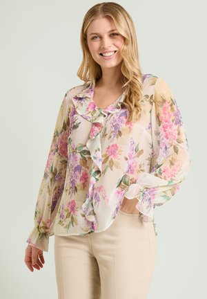 Glunderende vrouw met lang blond haar, gekleed in een lichte bloes met ruches en bloemenprint en beige broek, staand met één hand in haar zak.