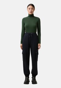 Vrouw die naar voren kijkt, gekleed in een groene coltrui, zwarte cargobroek met trekkoord in de taille en zwarte stevige schoenen op een witte achtergrond.