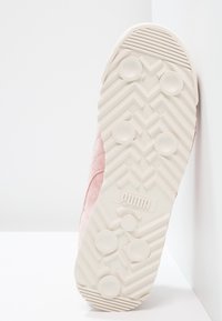 Světle růžové tenisky s texturovaným semišovým svrškem, bílé gumové podrážky s cik-cak vzorem a kruhovými protiskluzovými gripy, označeno "PUMA."