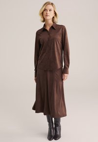 Brązowa sukienka koszulowa zamszowa z długimi rękawami, zapięciem na guziki z przodu i spódnicą o długości do połowy łydki. Połączona z czarnymi skórzanymi kozakami do kolan.