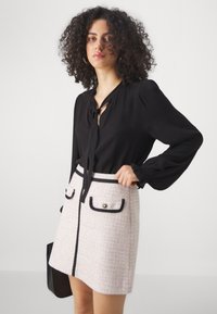 Blouse noire avec détails à nouer et manches longues ; jupe en tweed rose avec bord noir et poches contrastées ; texture lisse et forme structurée.