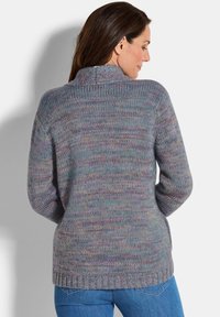 Femme aux longs cheveux bruns portant un pull en tricot multicolore et un jean bleu, se tenant de dos devant un mur uni.