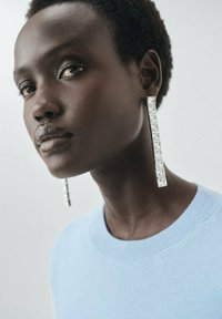 De longues boucles d'oreilles en argent avec des pierres rectangulaires transparentes, assorties à un pull bleu clair. L'encolure est ronde et texturée.