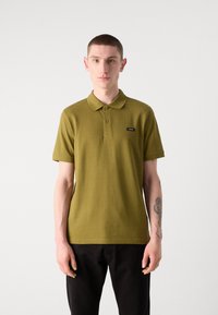 Camisa polo de color verde oliva con cuello, mangas cortas y un parche de logo negro. La tela parece suave; combinada con pantalones negros.