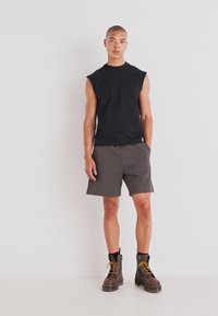 Zwarte mouwloze shirt gecombineerd met grijze shorts, gedragen met bruine veterschoenen met gele accenten. Eenvoudig, modern ontwerp met een relaxte pasvorm.