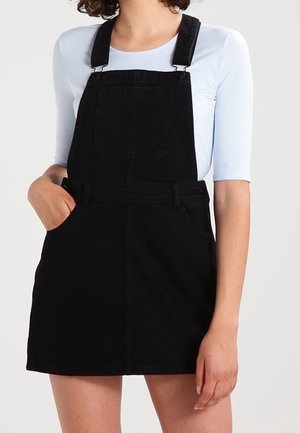Femme portant une robe salopette en jean noire avec une poche avant et un haut bleu clair à manches trois-quarts, main dans la poche.