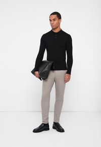 Sort, langermet polo genser med krage, beige bukser og svarte sko. Modellen holder en svart lærveske, og står mot en hvit bakgrunn.