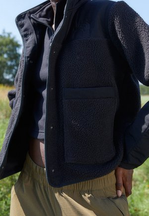 Persoon draagt een zwarte fleecejas met een grote zak over een zwarte trui met rits en kaki broek buiten in daglicht.