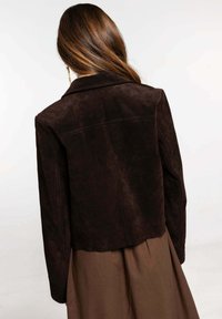 Chaqueta de ante marrón con un diseño cortado, cuello con muescas y detalles de costura visibles, combinada con una prenda marrón debajo.