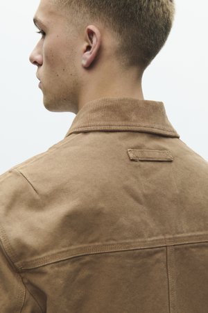 Jaunas vyras su trumpais blondinais plaukais dėvi smėlio spalvos drobinę striukę, matomas iš nugaros ir šiek tiek pasisukęs į kairę.