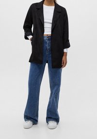 Blazer en daim noir avec larges revers, devant ouvert et manches retroussées, associé à un jean large pattes d'éléphant bleu taille haute et des baskets blanches.