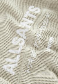 Tessuto beige dalla texture con testo ricamato bianco "ALLSAINTS" e ulteriore scrittura giapponese. Il design presenta un motivo a nido d'ape e accenti cuciti.