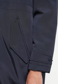 Cappotto impermeabile blu scuro con tasca laterale con patta, dettagli ai polsini con bottone e texture liscia. Design minimalista con linee pulite.