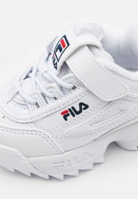 Sneaker in pelle bianca con strappo in velcro e lacci. Presenta il logo FILA in rosso e blu. Suola texturizzata con scanalature evidenti.