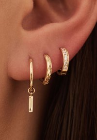 Drie gouden oorbellen: een gestructureerde ring, een gladde ring met een hangend rechthoekje, en een gebogen ring met vijf kleine heldere stenen.