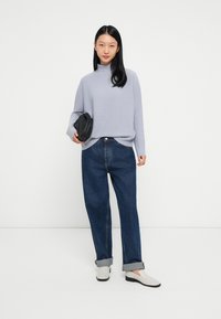 Žena stoji naslonjena na bijelu pozadinu, nosi svijetlosivi pulover s visokom ovratnikom, široke tamnoplave traperice s manžetama, bijele loaferice i drži crnu clutch torbicu.
