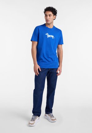 Modré bavlnené tričko s geometrickým motívom jazvečíka v bielej a modrej farbe. Kombinované s tmavými džínsami a béžovo-bielymi teniskami.