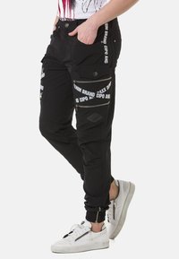 Schwarze Cargo-Hosen mit mehreren Reißverschluss- und aufgesetzten Taschen, versehen mit weißem Branding an der Seite. Das Material wirkt strapazierfähig und hat eine taillierte Passform.