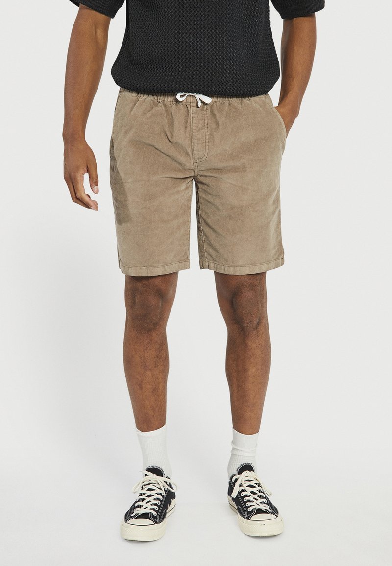 Άνδρας που φοράει μπεζ σορτς με κορδόνι στη μέση, λευκές κάλτσες, μαύρα και λευκά αθλητικά παπούτσια, και μαύρο κοντομάνικο πουκάμισο με υφή.