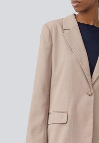 Beige blazer with a structured design, single button closure, notch lapels, and side pockets. Made from a smooth, woven fabric.