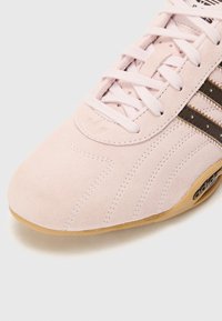 Lys rosa suedet sneaker med hvide snørebånd, tre brune striber og en tekstureret tåboks. Har en gummisål og et prægede logo.