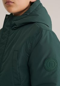 Tuvums kadrs ar cilvēku, kurš valk�ā tumši zaļu jaku ar augstu apkakli un apļveida logotipa uzšuvumu augšējā piedurknē.