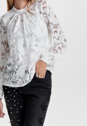 Frau trägt eine weiße Spitzenbluse mit Blumenmuster und langen Ärmeln, einem gerafften Ausschnitt und schwarze Jeans mit Perlen- und Spitzenapplikationen.