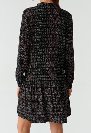 Zwarte jurk met lange mouwen, voorzien van een geometrisch patroon in rood en beige, textuurstof en een uitlopende zoom.