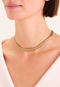 Frau mit einer kurzen, massiven Goldkette, die sie zu einem weißen Oberteil trägt, gezeigt von Kinn bis zur oberen Brust.