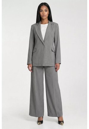 Kobieta z prostymi czarnymi włosami nosi szarą marynarkę w prążki i szerokie spodnie z czarnymi spiczastymi butami na białym tle.