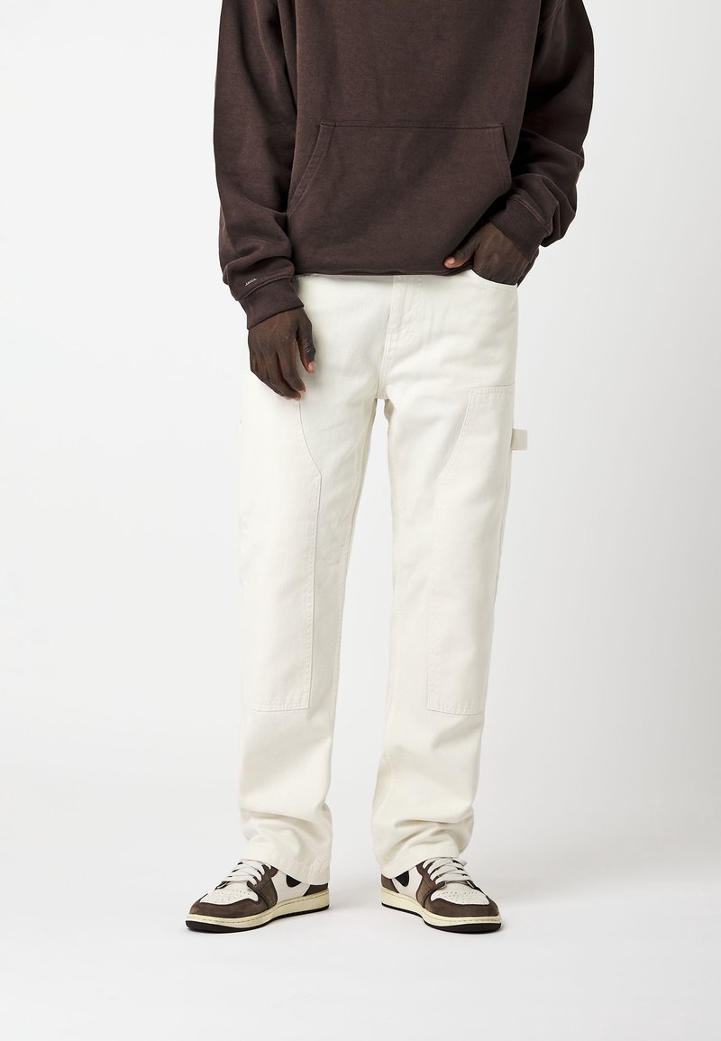 Personne portant un pantalon droit écru, des baskets blanches et marron, et un sweat à capuche marron foncé, debout devant un fond uni.