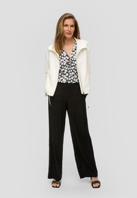 Witte zip-up jas over een zwart-wit geblokte blouse, gecombineerd met zwarte wijde broek en zwarte slide sandalen.