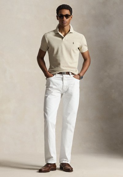 Man draagt een beige polo, witte broek, bruine riem en schoenen, donkere zonnebril, staat met handen in de zakken tegen een neutrale achtergrond.