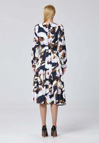 Une femme aux longs cheveux blonds se tient de dos, vêtue d'une robe fleurie à manches longues et arrivant au genou, avec des talons hauts noirs.