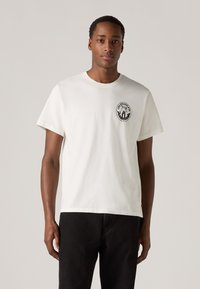 Hvit bomulls t-skjorte med rund hals, korte ermer og en svart logo grafikk på venstre bryst. Kombinert med mørke jeans.