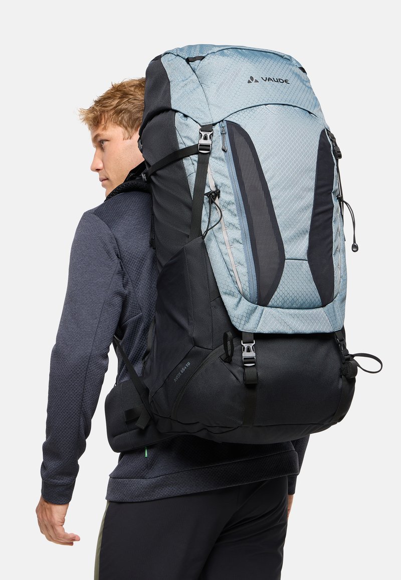 Homme portant un grand sac à dos de randonnée bleu et noir avec des bretelles rembourrées et plusieurs boucles, légèrement tourné vers la gauche.