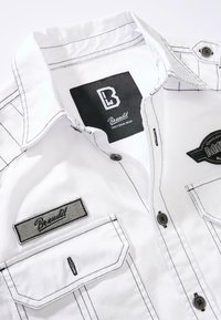White button-up shirt featuring contrast black stitching, two chest pockets, and embroidered brand labels on the left pocket and collar.