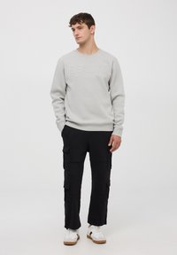 Felpa grigia chiara con testo in rilievo "REALITY" abbinata a pantaloni cargo neri con molte tasche. Sneakers bianche con suole marroni.