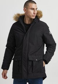 Sort polstret parka med pelsforet hætte, to forhåndsrum, lynlås og tekstureret detalje på brystet. Holdbart materiale, afslappet pasform.